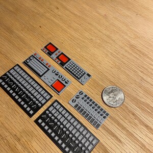 May include: A collection of small, grey and black 3D-printed control panels featuring different button and switch layouts. Some panels have red square details. A UK coin is included for scale. The items are displayed on a wooden surface.