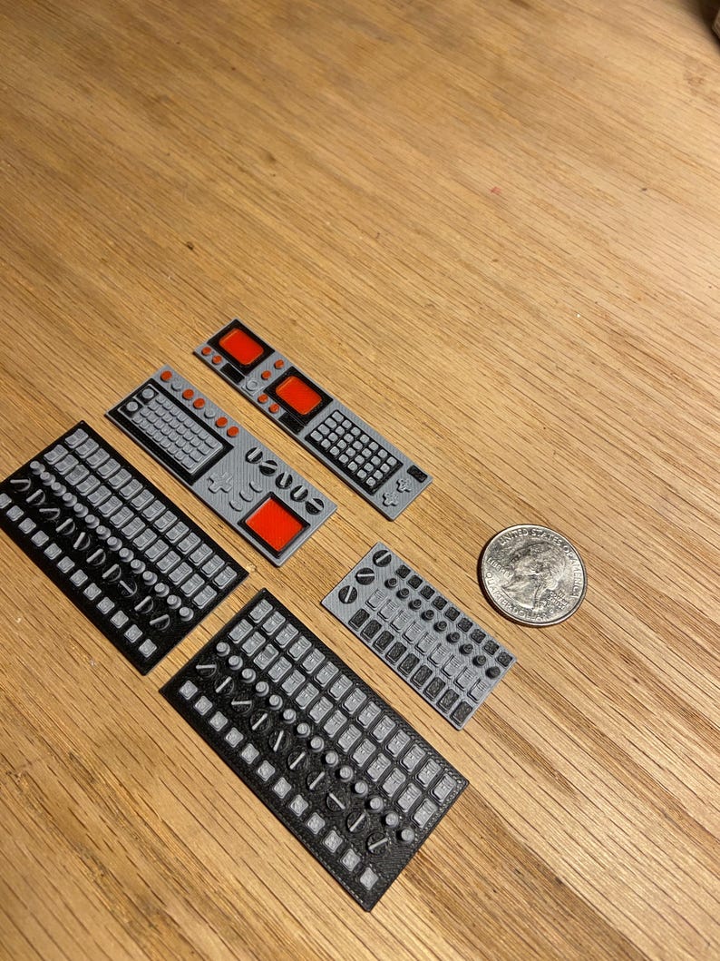 May include: A collection of small, grey and black 3D-printed control panels featuring different button and switch layouts. Some panels have red square details. A UK coin is included for scale. The items are displayed on a wooden surface.