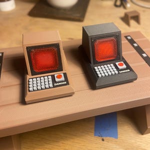 May include: Two miniature computer models, one tan and one black, featuring a bright red screen, a white grid keypad, and a small red button labeled DANGER. These 3D printed replicas sit on a tan surface with a textured, layered finish.