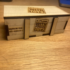 May include: A small, rectangular wooden box with laser-etched text and design. The box features the words "A PUNK WITH TOYS" and "WALK WITH TOYS". The box is made of light-colored wood and has a rustic appearance. The box has a lid and is held together with wooden supports.