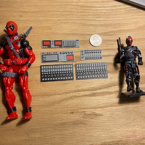 May include: Two Deadpool action figures, one in a red and black suit and the other in a grey and black tactical suit. Several small grey and black computer-like components are also present, along with a US quarter.