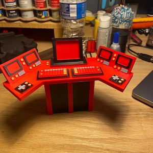 May include: A red and black miniature control panel with a central screen, keyboard, and multiple smaller screens. The model has a retro design with a black frame and red panels, and is likely a prop or toy.