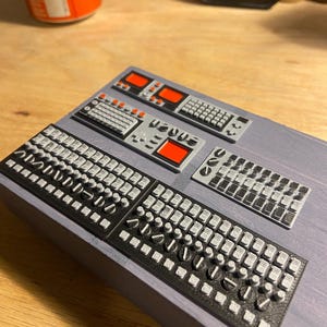 May include: Miniature electronic control panels with black and grey keyboards, buttons, and red display screens. The panels are arranged on a grey surface, suggesting a model or prototype of a control system.