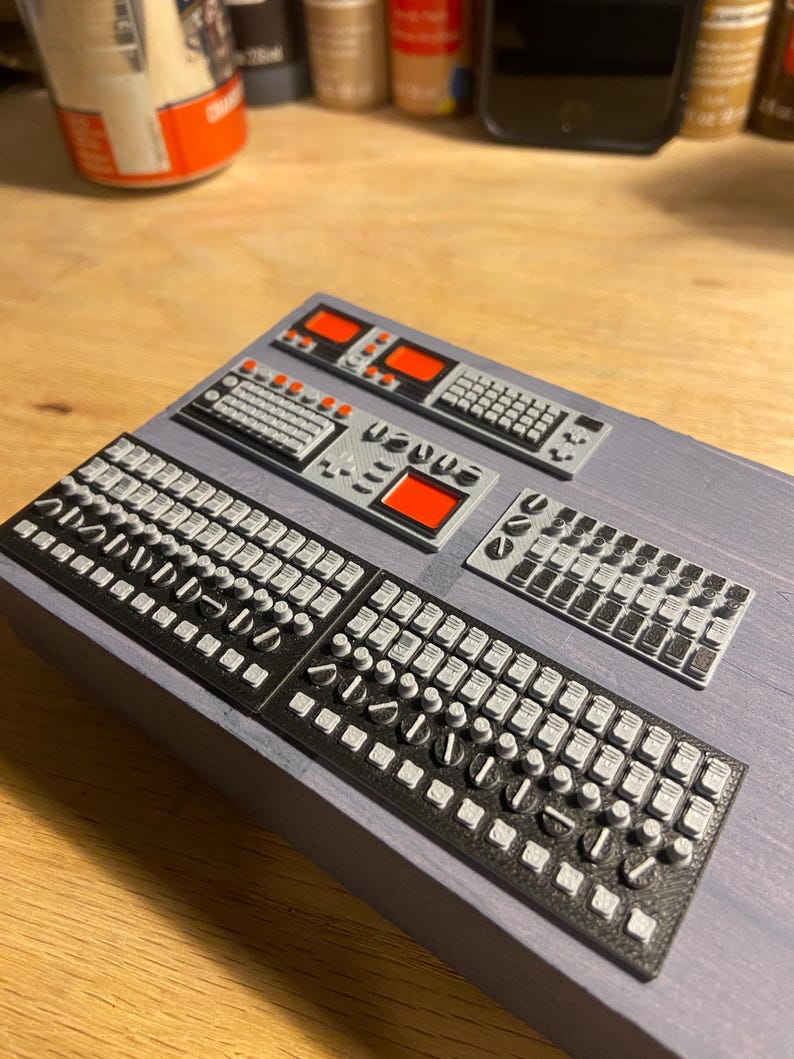 May include: Miniature electronic control panels with black and grey keyboards, buttons, and red display screens. The panels are arranged on a grey surface, suggesting a model or prototype of a control system.