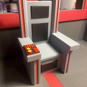 May include: A gray and red miniature chair with a control panel on the armrest. The chair has a rectangular back with two black squares and red accents. The armrest features a control panel with orange and yellow buttons.