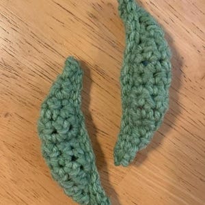 May include: Two handmade, green crochet leaf shapes. The leaves are made with a textured stitch, and are a soft green color. The leaves are lying on a wooden surface.