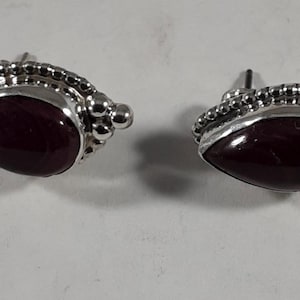 May include: A pair of stud earrings featuring a teardrop-shaped dark burgundy stone set in a silver frame. The silver setting is decorated with a beaded border and small silver spheres at one end, with a standard post back for secure attachment.
