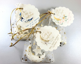 May include: White favor bags with gold stars and a white moon and star charm. The bags have tags that say "We're Over the Moon" with a blue and white moon and floral design. One tag says "Baby Gabriel Arriving in September 2023".