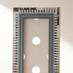 May include: A white rectangular crafting tool featuring the text PERFECT SLIMLINE LAYERS and MINI SLIMLINE. The tool has a central rectangular cutout with four circular holes, surrounded by blue and gray borders and numbered scales along the edges.