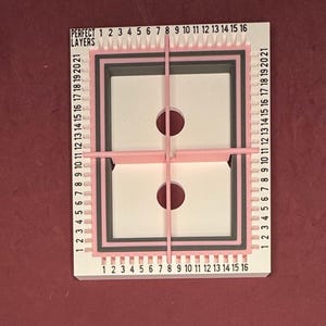 May include: A white and pink layered crafting tool with a rectangular design. The tool has a series of numbered markings and the text "PERFECT LAYERS". Two circular cutouts are visible within the central white space. The tool is designed for precise layering.