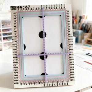 May include: A white rectangular crafting alignment tool featuring black printed numbers and text reading PROJECT LAYERS A6-A7. It includes light blue and pink nested frames and two perpendicular lavender notched bars for precise positioning.