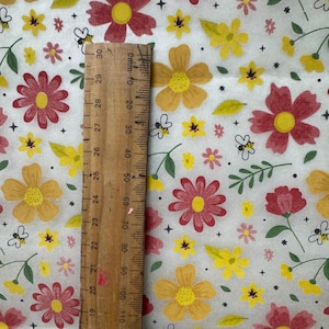 May include: A fabric with a floral pattern featuring red, yellow, and pink flowers, green leaves, and small bees on a white background. A wooden ruler with centimeter markings is placed on the fabric, showing measurements up to 30 cm.