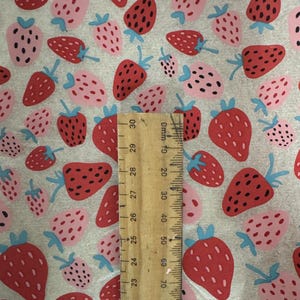 Pode incluir: Um tecido com um padrão repetido de morangos vermelhos e rosa com caules verdes e folhas azuis. Uma régua de madeira é colocada sobre o tecido, mostrando medidas em milímetros e centímetros.