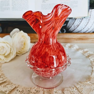 May include: A vibrant red glass vase with a ruffled top and a round base. The vase has a speckled pattern and sits on a clear, circular foot. The vase is a decorative item.