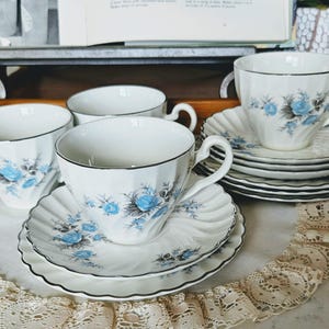 May include: A set of white teacups and saucers with a blue floral design and black trim. The teacups have a classic shape and are arranged on a white surface with a lace trim. The set includes multiple cups and saucers.