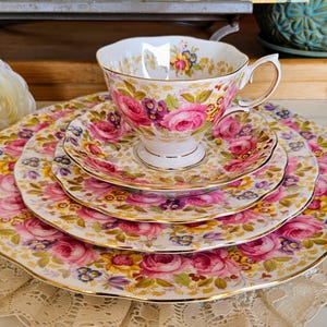 Puede incluir: Juego de té vintage con un diseño de rosas florales en rosa, morado, amarillo y azul. El juego incluye una taza de té, un platillo y platos con ribetes dorados y borde festoneado. Un conjunto de vajilla elegante.