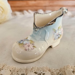 May include: A small ceramic shoe-shaped figurine with a cream-colored base, featuring hand-painted light blue and pink floral motifs. The rim of the shoe is accented with a metallic silver trim, and the overall finish is glossy and smooth.