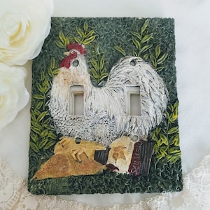 以下が含まれることがあります： 緑と茶色のセラミック製のライトスイッチカバーに、赤い鶏冠と赤い肉垂れを持つ白い雄鶏が描かれています。雄鶏は緑の葉と茶色のテクスチャのある背景に囲まれています。スイッチカバーには、ライトスイッチ用の開口部が2つあります。