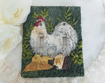 Può includere: Una placca per interruttore in ceramica verde e marrone con un gallo bianco con cresta e bargiglio rossi. Il gallo è circondato da foglie verdi e uno sfondo marrone testurizzato. La placca ha due aperture per interruttori.