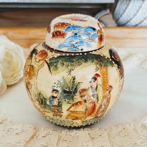 May include: A round ceramic jar with a lid, featuring a detailed painted scene. The jar's design includes figures in a landscape, with green, brown, and gold hues. The lid is decorated with blue waves and red accents.