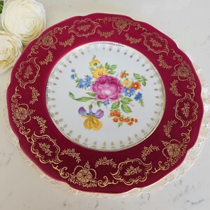 May include: A decorative plate with a claret-coloured rim and a floral design. The rim features gold filigree patterns. The centre of the plate displays a bouquet of colourful flowers, including pink roses, blue forget-me-nots, and yellow irises.