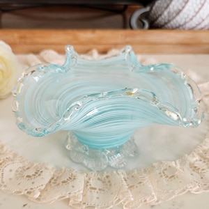 Puede incluir: Un cuenco de cristal azul claro con borde ondulado y base de pedestal. El cuenco tiene un diseño en espiral y se asienta sobre un tapete de encaje blanco. Es un objeto decorativo.
