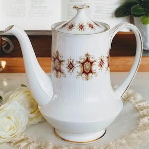 Puede incluir: Tetera de cerámica blanca con ribete dorado y diseño floral rojo y dorado. La tetera tiene un pico y una asa.