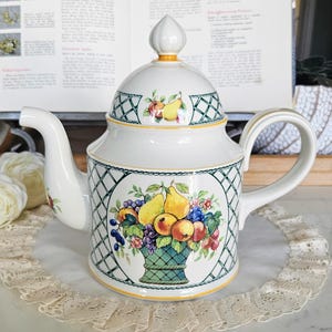 May include: White ceramic teapot with a lid, featuring a fruit basket design. The teapot has a curved spout and handle, with a green lattice pattern and yellow accents. The fruit basket includes pears, grapes, and apples.