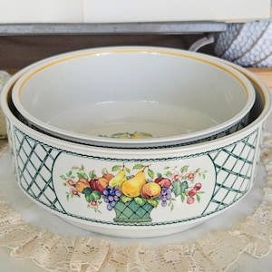 Puede incluir: Dos cuencos de cerámica blanca. El cuenco más grande tiene un diseño de enrejado verde con una cesta de frutas. El cuenco más pequeño está anidado dentro. Los bordes tienen un ribete amarillo y negro. Los cuencos están sobre un mantel individual de encaje.