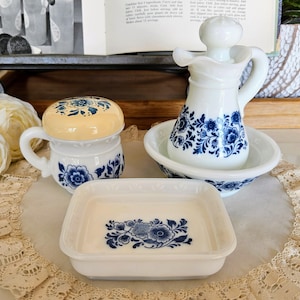Könnte beinhalten: Eine Sammlung weißer Keramik-Küchenutensilien mit blauen Blumenmustern. Das Set umfasst einen kleinen Becher mit gelbem Deckel, einen Krug mit einer Schüssel und eine rechteckige Schale. Die Teile sind auf einer Spitzendecke angeordnet.
