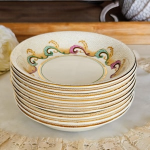 May include: A stack of vintage porcelain saucers. Each saucer has a cream-colored background with a gold rim and a decorative border featuring teal, pink, and gold accents. The saucers are stacked on a white lace doily.