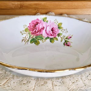 May include: Oval white porcelain serving dish with a gold rim and a floral rose design. The dish features two pink roses with green leaves and small pink buds. The dish is placed on a white lace doily.