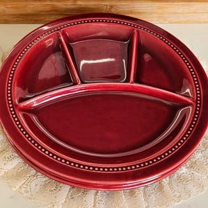 May include: A deep red, divided serving platter with a glossy finish. The round plate has three sections and a dotted border. The plate is sitting on a white lace doily.