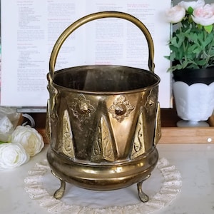 Peut inclure: Un chaudron en laiton antique avec une poignée, reposant sur trois petits pieds. Le chaudron présente des motifs décoratifs en forme d'étoiles et de triangles autour du bord. L'intérieur est sombre et le laiton a une patine chaude et vieillie. Un livre de cuisine vintage est en arrière-plan.