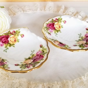 Puede incluir: Dos platos blancos en forma de concha con borde dorado. Cada plato está decorado con un motivo floral de rosas rojas, amarillas y rosas. Los platos están sobre una superficie blanca con encaje.