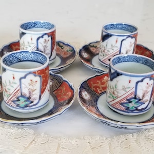 May include: Set of four Japanese-style tea cups and saucers. The cups and saucers feature a white base with intricate designs in red, blue, and gold. The cups are cylindrical, and the saucers are oval-shaped.