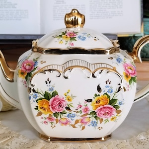 Könnte beinhalten: Eine weiße Teekanne mit goldener Verzierung und Blumenmuster. Die Teekanne hat einen Deckel mit goldenem Knauf und einen Henkel mit goldener Verzierung. Die Teekanne ist mit rosa, gelben und blauen Blumen verziert.