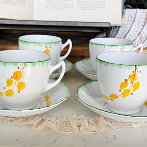 Puede incluir: Cuatro tazas y platillos blancos con borde verde y diseño floral amarillo. Las tazas tienen un asa curva y están sobre una superficie blanca. Los platillos tienen un borde verde a juego.