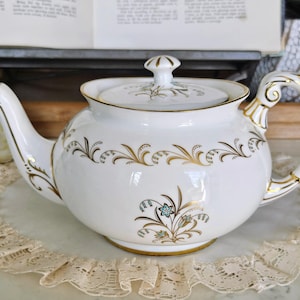May include: White porcelain teapot with gold trim and floral design. The teapot has a lid with a finial and a curved handle. The floral design includes gold and teal accents. The teapot is on a lace doily.