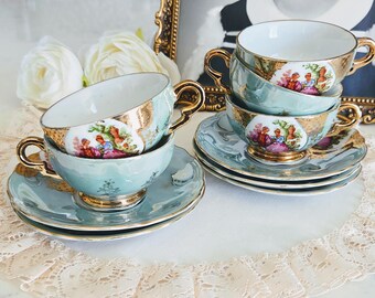 Puede incluir: Un juego de cuatro tazas de té y platillos de porcelana azul claro con asas y bordes dorados. Cada taza presenta un medallón circular central con una ilustración colorida de dos figuras en un jardín, acentuado con detalles dorados.