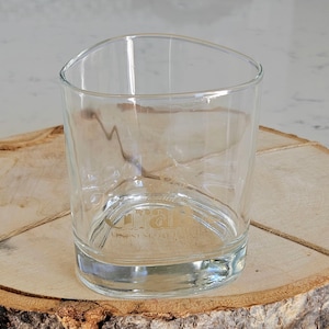 May include: A clear, triangular-shaped glass tumbler with a thick base. The glass sits on a round, light brown wooden slice with a natural bark edge. The glass has text etched on the bottom.