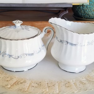Könnte beinhalten: Weißes Porzellan-Zucker- und Sahne-Set. Beide Teile haben einen silbernen Rand und ein zartes blaues Blumendesign. Die Zuckerdose hat einen Deckel mit dekorativem Knauf. Die Sahnekännchen hat einen gebogenen Griff und Ausguss. Maße in cm.
