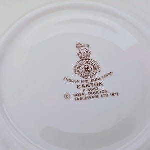 May include: A white plate featuring the Royal Doulton Canton logo in brown, with text including "English Fine Bone China" and "Tableware Ltd. 1977". The plate has a curved rim.