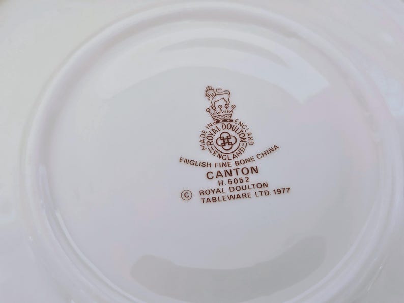 May include: A white plate featuring the Royal Doulton Canton logo in brown, with text including "English Fine Bone China" and "Tableware Ltd. 1977". The plate has a curved rim.