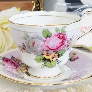 Puede incluir: Una taza de té y un platillo blancos con un diseño floral. La taza de té tiene una rosa rosa y blanca con hojas verdes y flores amarillas. El platillo es de color morado claro con un borde dorado.