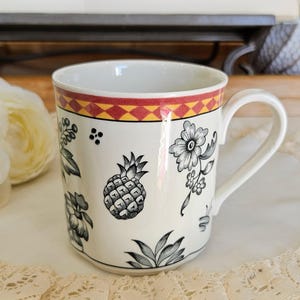 Puede incluir: Taza de cerámica blanca con una banda decorativa de formas geométricas rojas y amarillas en el borde. La taza presenta ilustraciones florales y de piña en blanco y negro. Un asa curva está unida al lateral.