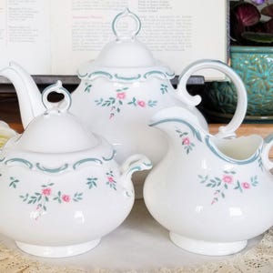Puede incluir: Un juego de té de porcelana blanca con tetera, azucarero y jarra de crema. Cada pieza está decorada con ramitas florales rosas y un ribete festoneado azul claro. El juego se exhibe sobre un tapete de encaje.