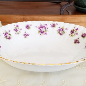May include: White oval serving bowl with a scalloped edge and gold trim. The bowl is decorated with clusters of purple and green floral designs. A decorative piece for serving food.