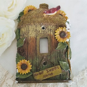 Puede incluir: Una cubierta decorativa para interruptor de luz con forma de casa de pájaros rústica. La cubierta presenta un tejado de paja, girasoles y un pequeño pájaro. Las palabras "Home Sweet Home" están en un pequeño letrero. La paleta de colores incluye tonos marrones, verdes y amarillos.