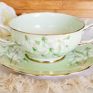 May include: A light green teacup and saucer set with a floral design. The teacup has two handles and a gold rim. The saucer has a scalloped edge and matches the teacup's design. The set is decorated with white and green floral patterns.